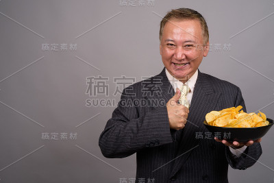 灰色背景下，一名成熟的亚洲商人捧着一碗薯片