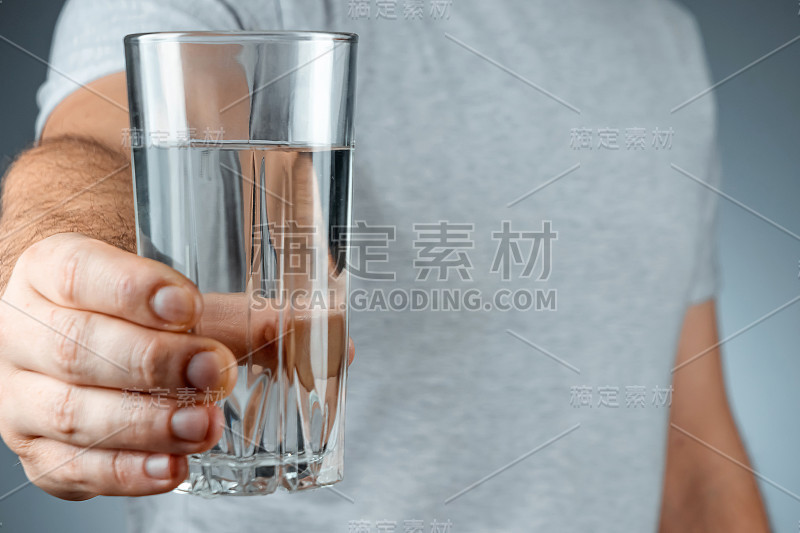 玻璃杯与纯矿泉水在男性的手特写在一个灰色的背景。药物治疗健康饮品