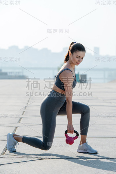 一个漂亮的褐发女人带着小壶铃在户外的人行道上锻炼
