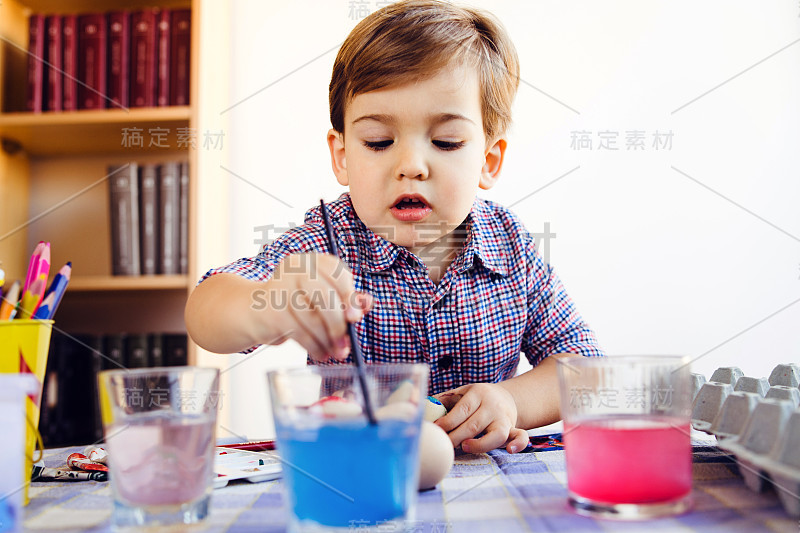 一个白人小男孩在家里的桌子边拿着蘸水的毛笔玩着水彩画复活节彩蛋