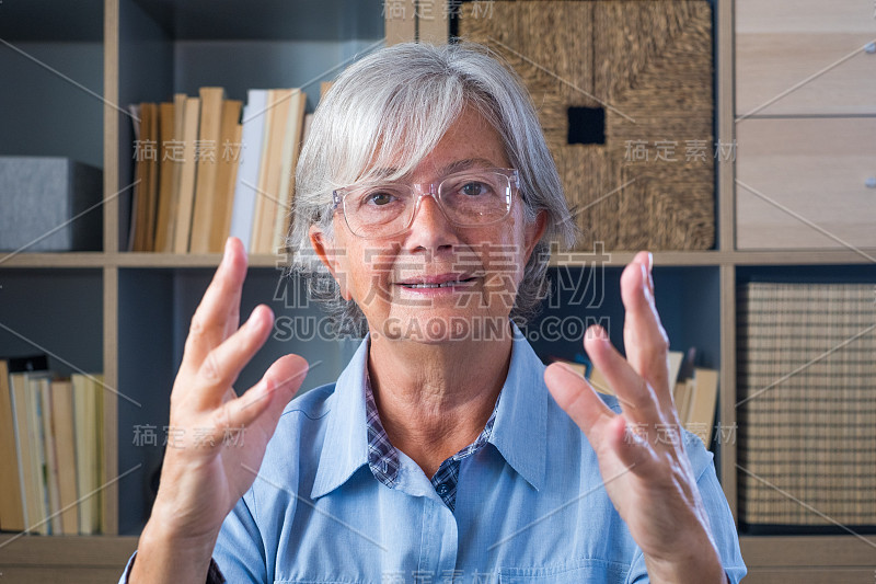 头像自信的老女商人教练戴着眼镜看着相机和说话，导师演讲者举行在线课程，解释，坐在现代橱柜的木制办公桌