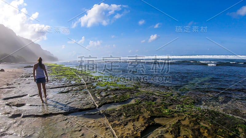 Badung -一个女人走在石头海滩上，覆盖着海藻和海藻