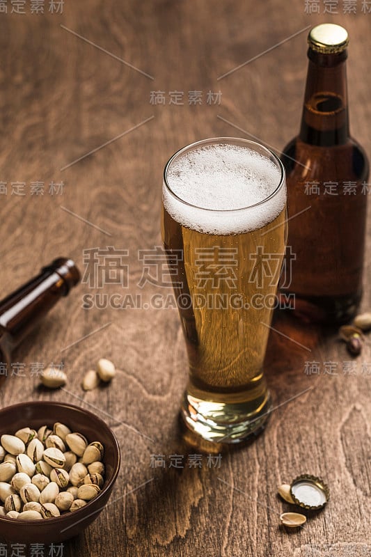 两个深色啤酒瓶，一杯啤酒和泡沫，一盘木色背景上的开心果。