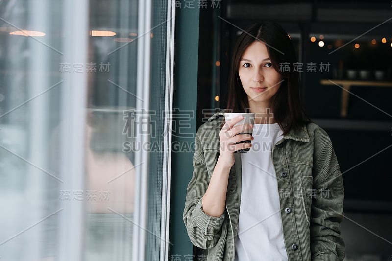 有魅力的女人站在窗边的卧室里，手里拿着马克杯