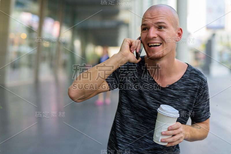 有思想的年轻快乐的秃顶男人微笑和大笑，在泰国曼谷的购物中心外打电话和拿着纸咖啡杯