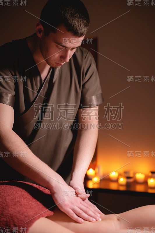 男士优质豪华臀部按摩减肥健康中心。男按摩师在办公室里给一位女客户做腿部按摩，昏暗的灯光映衬着燃烧的蜡