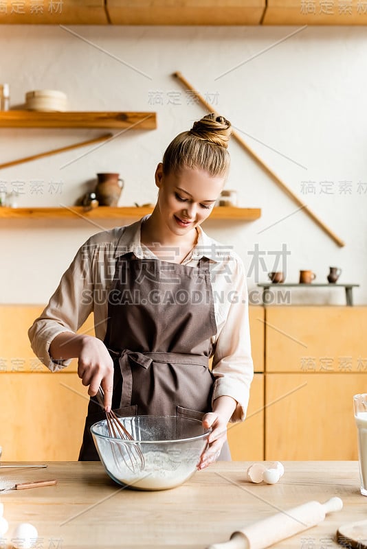 幸福的女人在围裙拿搅拌器附近的碗与面粉