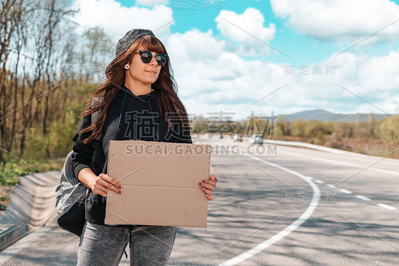 搭便车。微笑的年轻女子戴着太阳镜和帽子举着一个纸板标志与模拟。副本的空间。本地旅游的概念