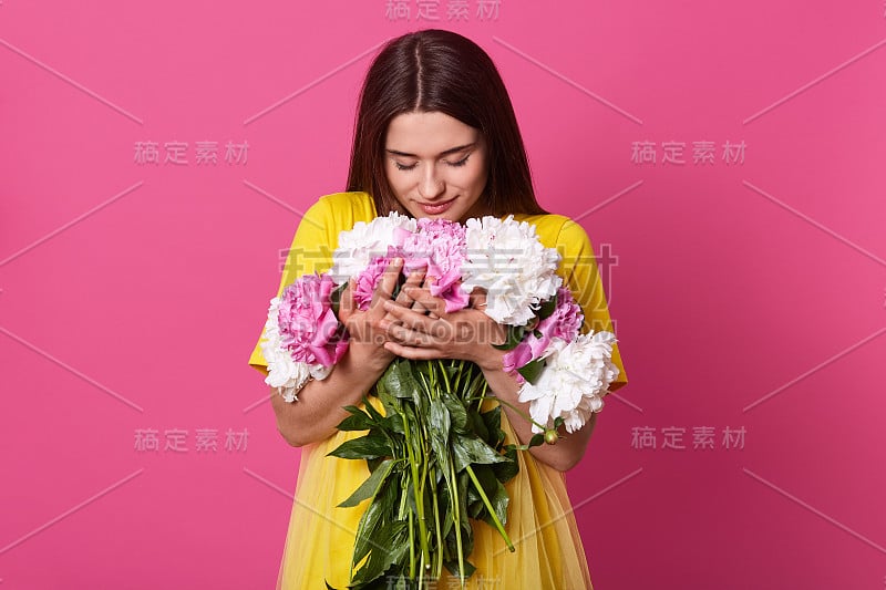 近距离的美丽的黑发女人拿着花孤立在粉红色的工作室背景，女士穿着黄色连衣裙拥抱大花束，女孩看花。3月8