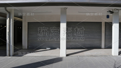 商店关上了卷帘门，放在有柱子的门廊下。白色的墙壁，阴影和前面的瓷砖路面。
