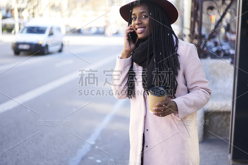 开朗的年轻的衣着漂亮的非洲裔美国女学生站在城市设置的大道上打出租车去大学，而有电话与服务订车在早上