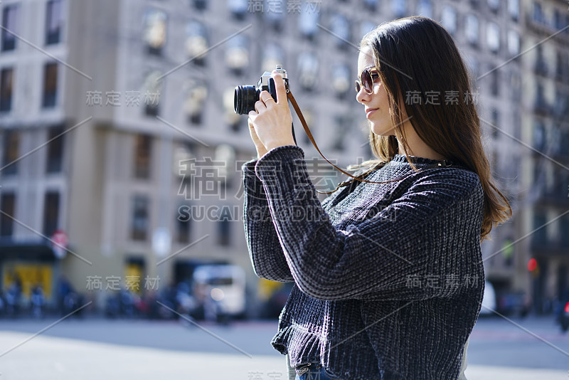 女旅行者戴着太阳镜拿着老式相机从周末观光旅游拍照。专业摄影师在阳光明媚的日子拍摄城市背景建筑