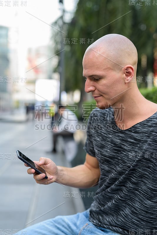 在泰国曼谷购物中心外使用手机的年轻英俊的秃头男人的侧面视图