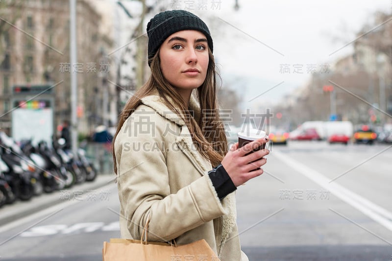 一名年轻的欧洲女子走在城市街道上，手里拿着外卖的咖啡