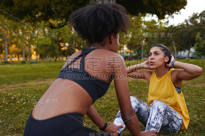 健康年轻女子做仰卧起坐与她的锻炼女性伙伴在绿色草地上锻炼期间-年轻决心和积极的妇女做运动