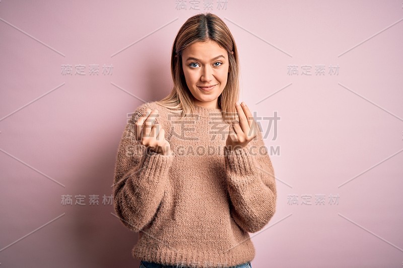 年轻美丽的金发女人穿着冬季羊毛毛衣在粉红色孤立的背景做钱手势与手，要求工资支付，百万富翁的业务