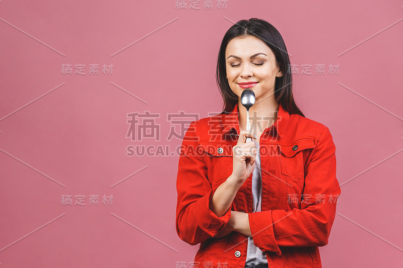 吃晚饭了!年轻美丽的女人的肖像穿着红色休闲衬衫，拿着勺子孤立在粉红色的背景。