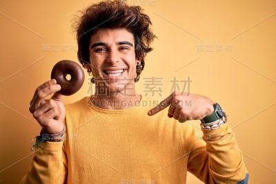 年轻英俊的男子拿着巧克力甜甜圈站在孤立的黄色背景与惊讶的脸指向自己
