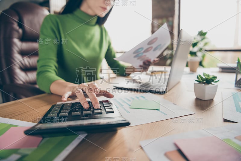 忙碌的商务女士的特写照片笔记本桌子拿着纸统计数字计算金钱收入坐椅子穿绿色高领现代办公室