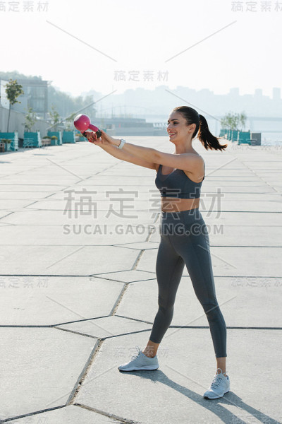 一个快乐的褐发女人带着小壶铃在户外的人行道上锻炼