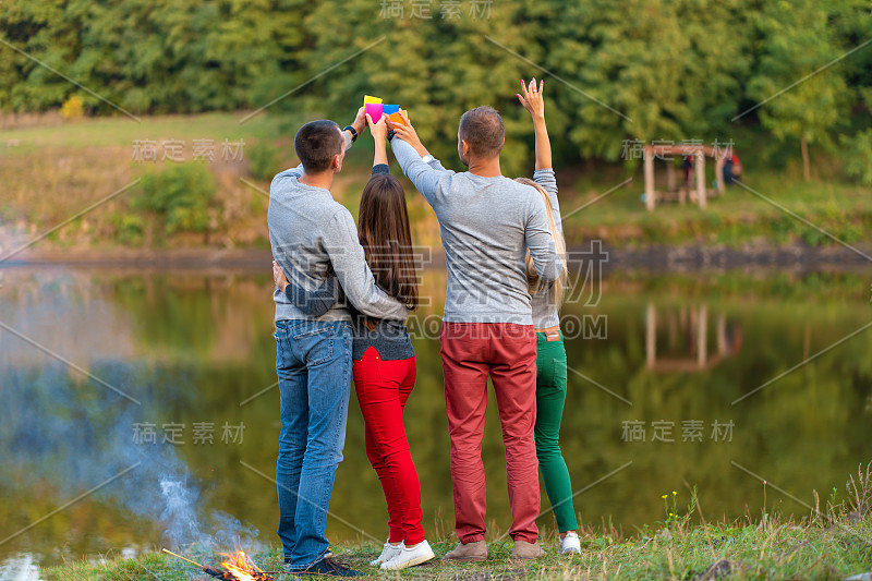 和朋友们在篝火附近的湖边野餐。公司朋友有徒步野餐的自然背景。徒步旅行者在喝酒时间放松。夏季野餐。与朋