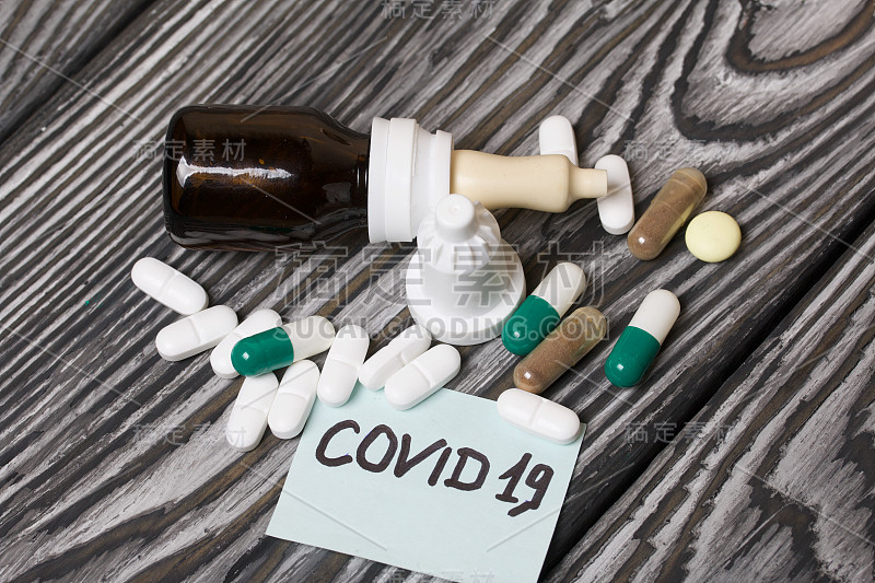 贴有COVID-19字样的贴纸。旁边是几颗形状和颜色各异的药丸。装有鼻腔喷雾剂的罐子。躺在刷过的松木