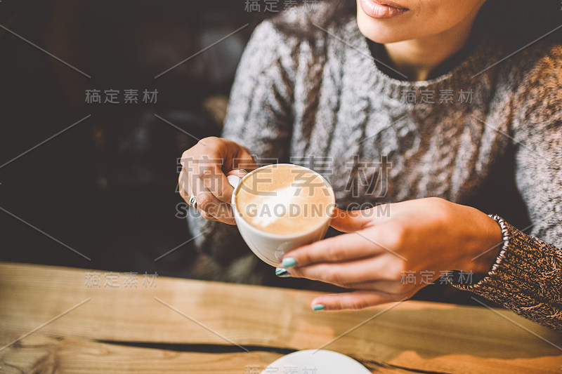 美丽的商务女孩在装饰有圣诞装饰的餐厅喝着白色的咖啡。靠窗的坐在木桌旁靠窗的穿着暖和的灰色毛衣。在桌子