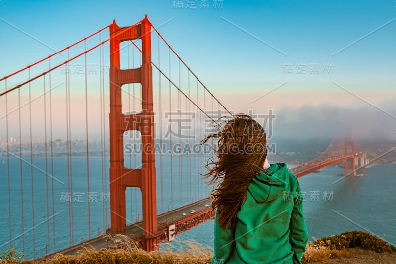 一名身穿绿色连帽衫的年轻女子站在一座山上，在日落时分俯瞰金门大桥，旧金山