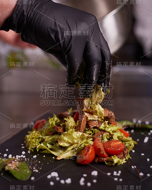 用小牛肉烹饪热沙拉的过程。一个戴着黑手套的厨师的手。黑色板岩板。