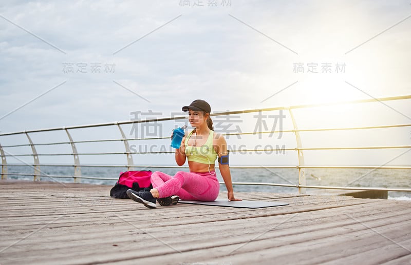 年轻健康的女人穿着运动服坐在垫子上，在海滩上喝水