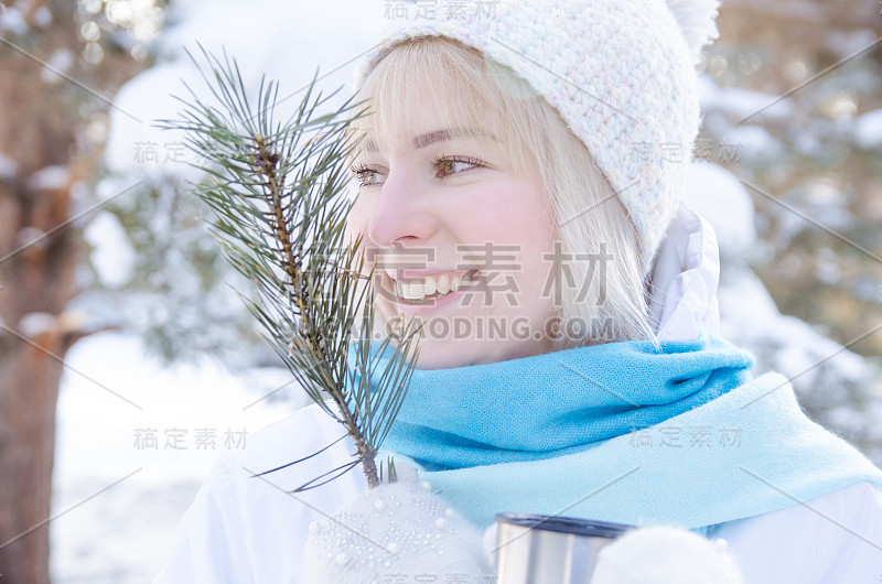 一个迷人的金发微笑女孩的肖像与云杉树枝和一个马克杯在一个雪山的森林里。