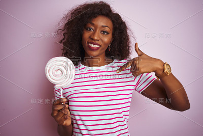 年轻的非洲裔美国妇女吃糖果站在孤立的粉红色背景与惊讶的脸指向自己的手指