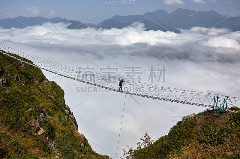 人们走在吊桥上，看着下面多云的山。