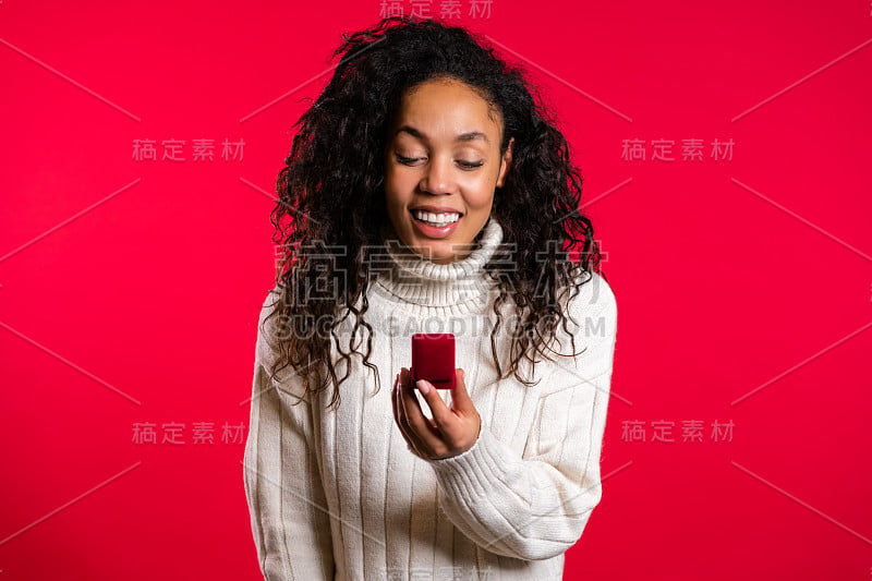 惊奇的非洲裔美国年轻女子拿着小首饰盒与求婚钻戒在红墙的背景。女孩微笑着，她高兴地得到礼物，求婚。