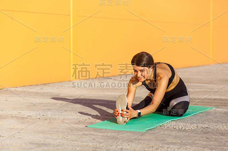 年轻美丽的运动瑜伽女性穿着黑色运动服伸展在街道地板上，坐在垫子上，直视