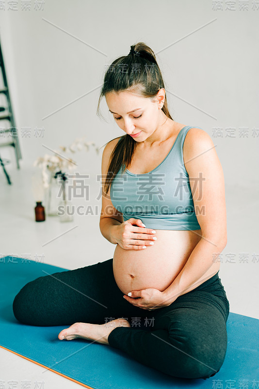 穿着运动服的漂亮怀孕女孩坐在瑜伽垫上，在健身房里抚摸着自己的肚子