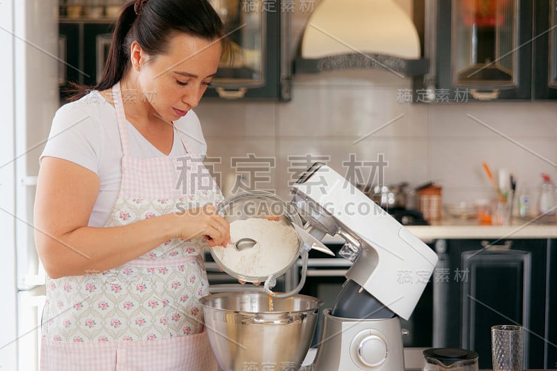 一位妇女在食品加工机的碗里加入面粉，准备烘焙。