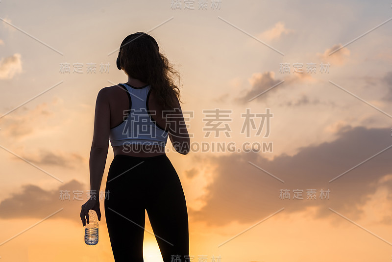 剪影年轻女子在耳机在运动服与一瓶清澈的矿泉水在日落天空的背景。健康生活方式的概念