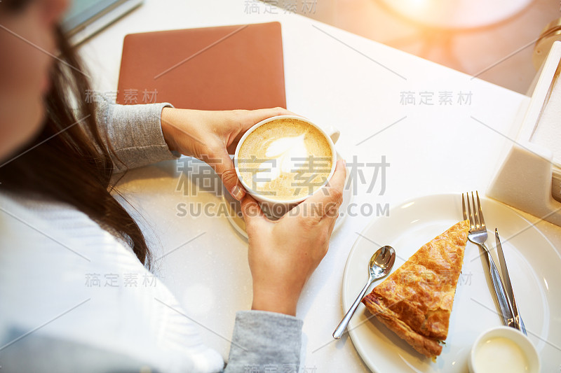 年轻微笑的女人做一个咖啡休息坐在咖啡馆，笔记本和一杯咖啡或拿铁，菠菜派，俯瞰