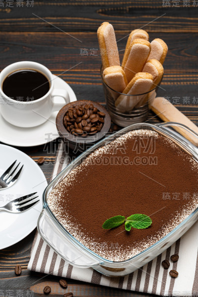 传统的意大利提拉米苏甜点在玻璃烤盘，savoiardi饼干和一杯新鲜的热咖啡在木背景