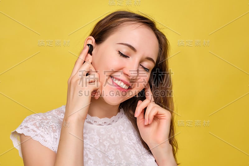 一个穿着夏装的女孩站在黄色的背景上，用手指拿着无线耳机，闭着眼睛听着耳机跟着唱。