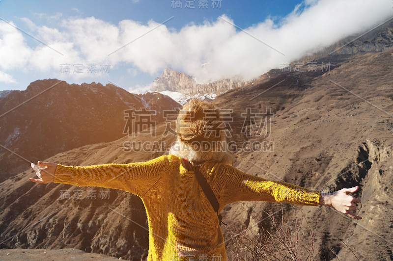 一个戴着大皮帽，穿着黄色针织衫的年轻女旅人，正张开双臂站在山中