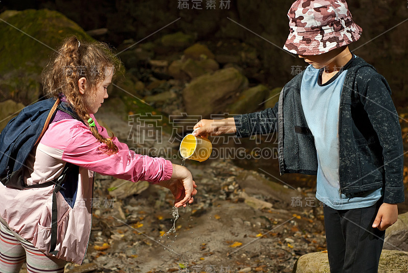 穿粉红色夹克的女孩在洗手。一个男孩从泉水旁的杯子里倒水。孩子们互相帮助洗手。援助和相互支持的概念。