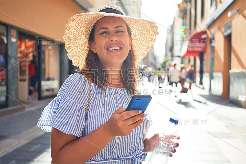 在一个阳光明媚的夏天，年轻美丽的女人用智能手机在城市的街道上快乐地走着