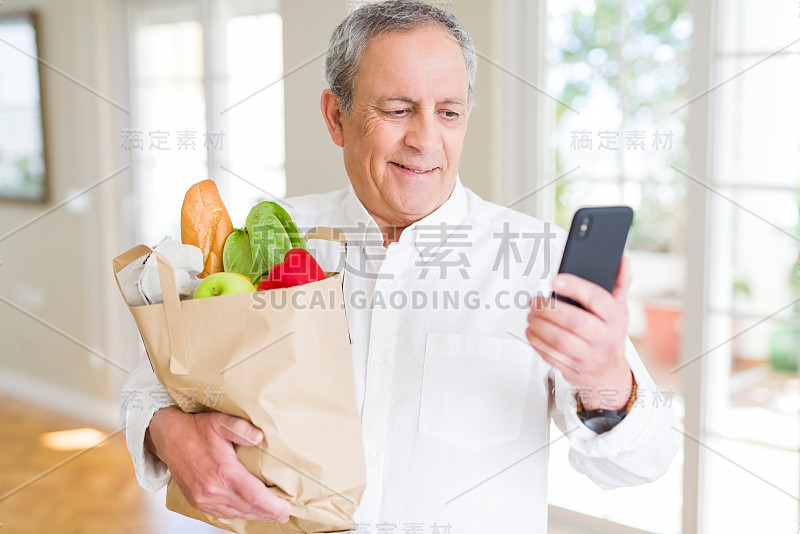 英俊的老男人拿着装满新鲜食品的纸袋，看着智能手机微笑