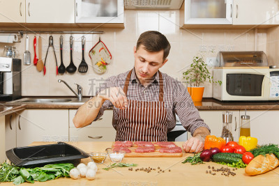 白种年轻人围着围裙坐在蔬菜桌前，在家做饭准备猪肉，牛肉或羊肉，盐肉，在明亮的厨房与木制表面，充满了花