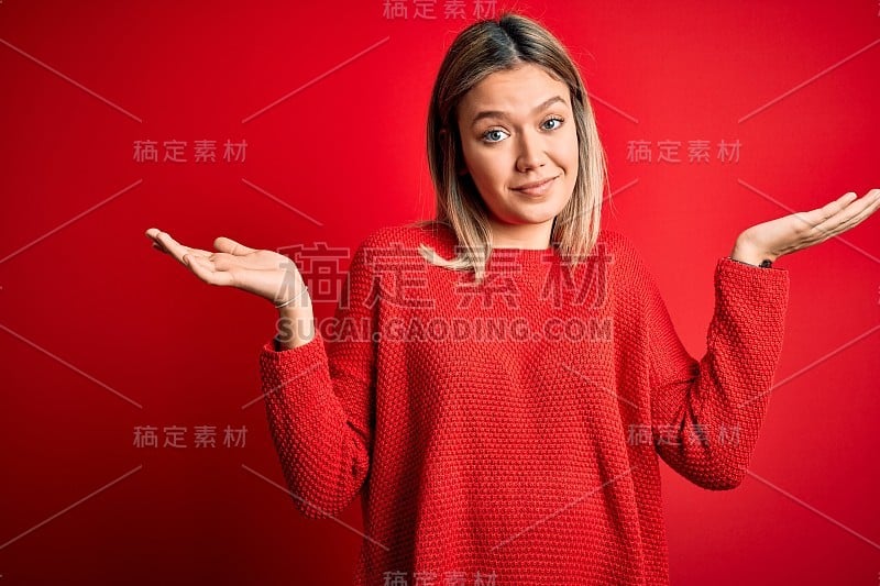 年轻美丽的金发女人穿着休闲毛衣在红色孤立的背景，毫无疑问和困惑的表情与胳膊和手举起。怀疑的概念。