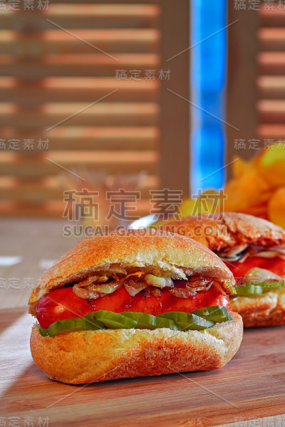 烤热狗配炸洋葱和腌黄瓜。薯片。木桌上的快餐。明亮的日光。