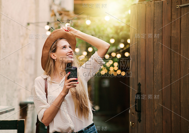 年轻迷人的女人在休闲风格的衣服走在老城的街道上，一杯咖啡在路上