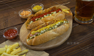 木桌上放着两个热狗，薯条，香料和大杯啤酒
